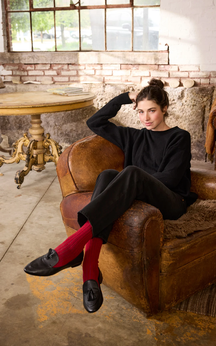 Woman wearing black sweatsuit with red ribbed socks and black leather loafers, sitting in vintage leather chair
