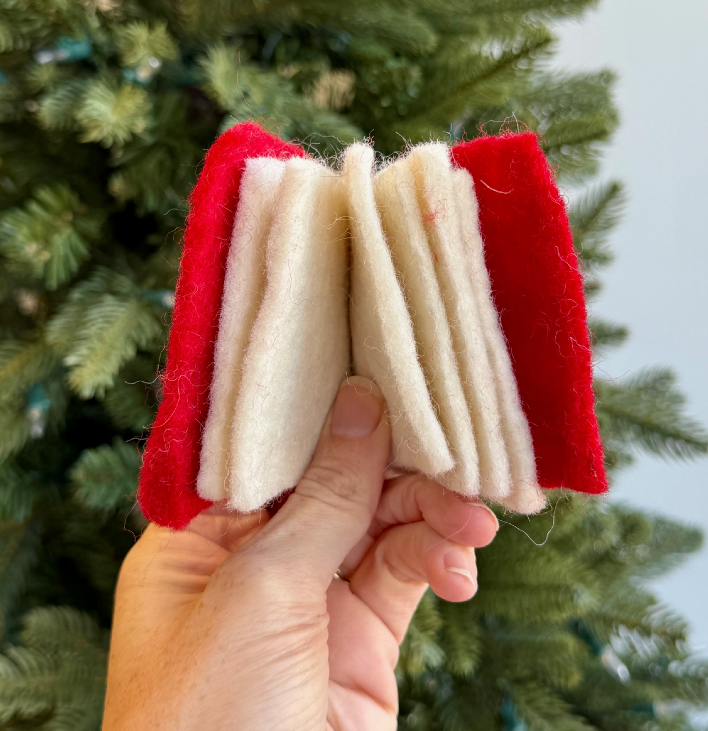 Handmade felt Santa beard ornament in red and cream, featuring accordion-folded white beard held against Christmas tree background