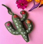 Green felt cactus ornament with white cross-stitch detail and colorful embroidered flowers on pink background