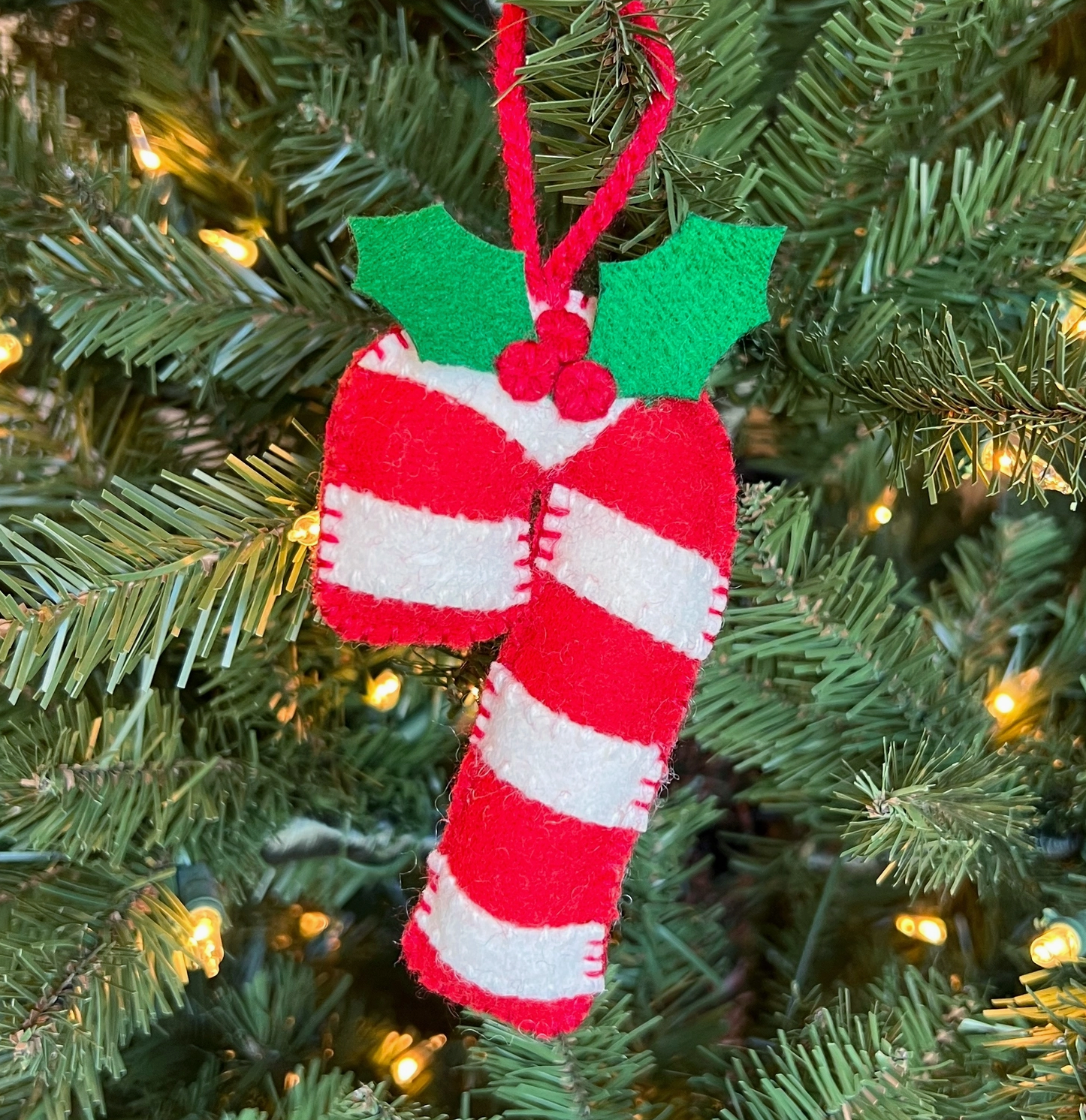 Handmade felt candy cane ornament with red and white stripes, green holly leaves, and red ribbon hanger on Christmas tree