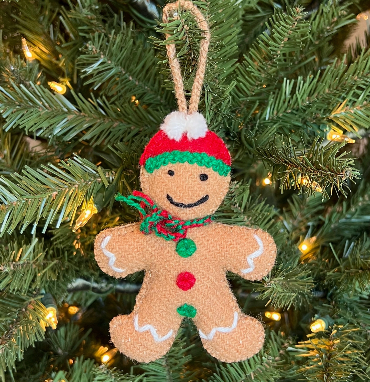 Handmade felt gingerbread man ornament with red and green hat, striped scarf, button details, and rope hanger on Christmas tree
