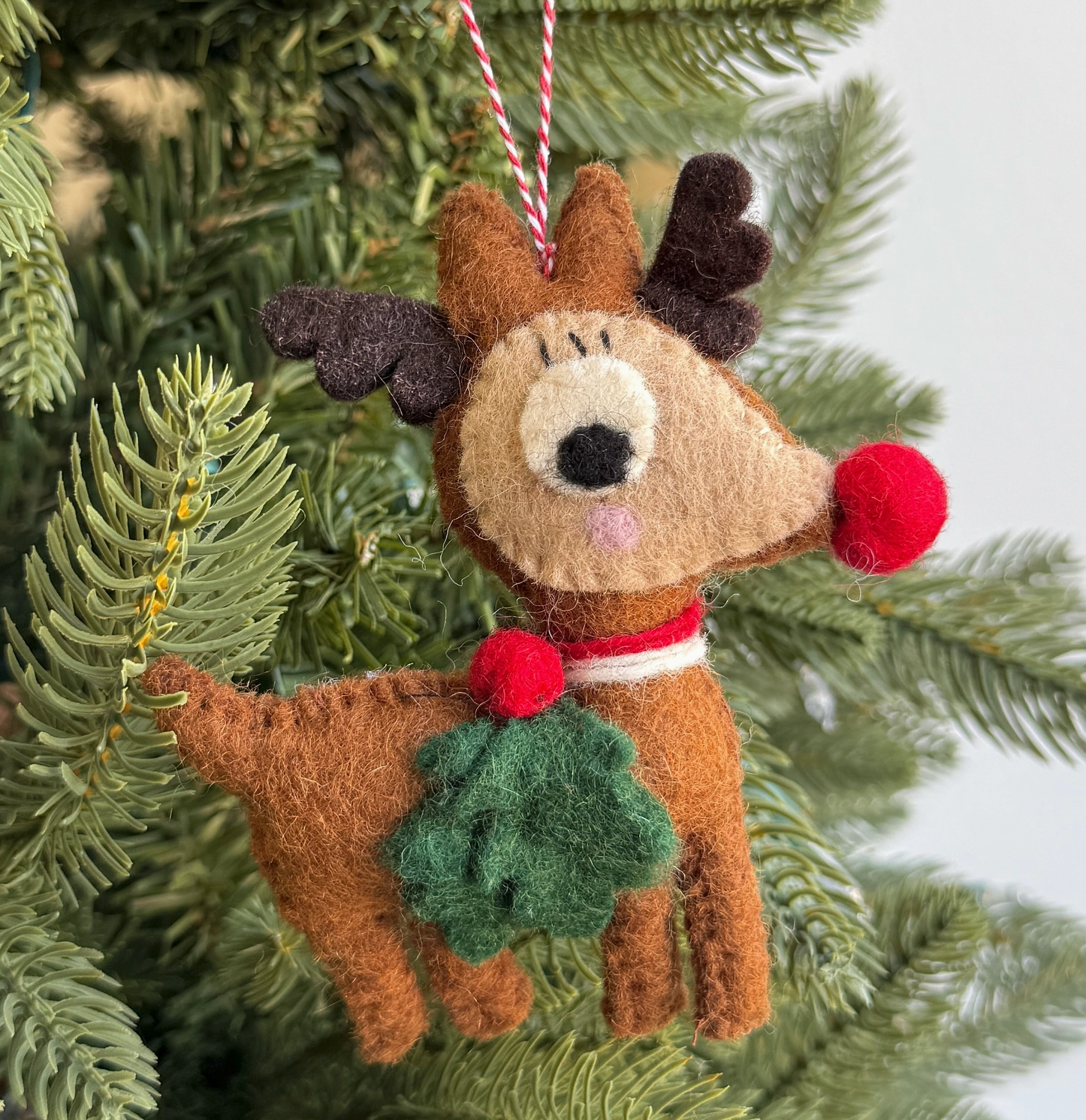 Handmade felt reindeer ornament in brown with red nose, antlers, collar and green wreath detail hanging on Christmas tree