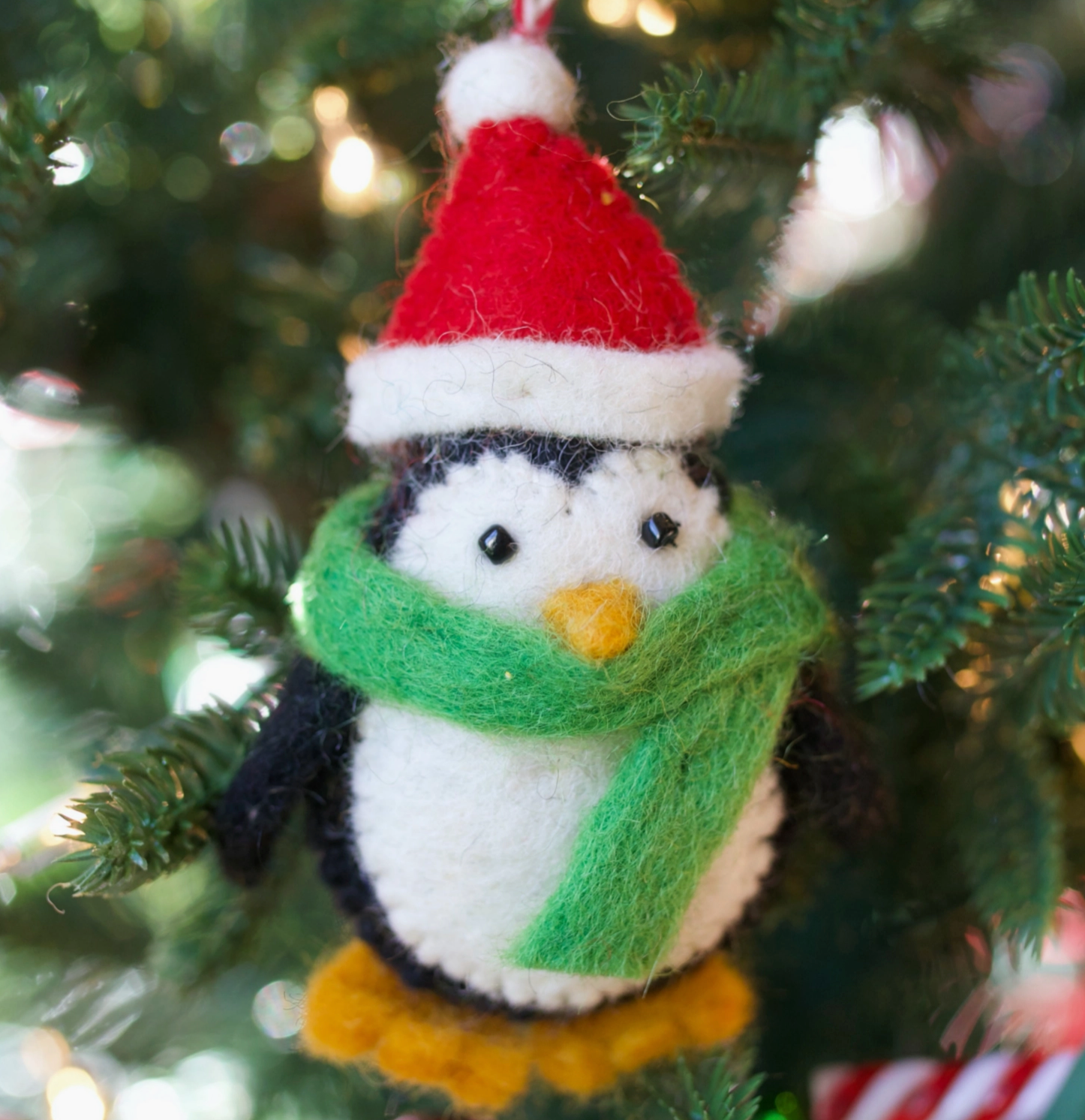 Felt penguin ornament with red Santa hat, green scarf, and orange beak hanging on Christmas tree