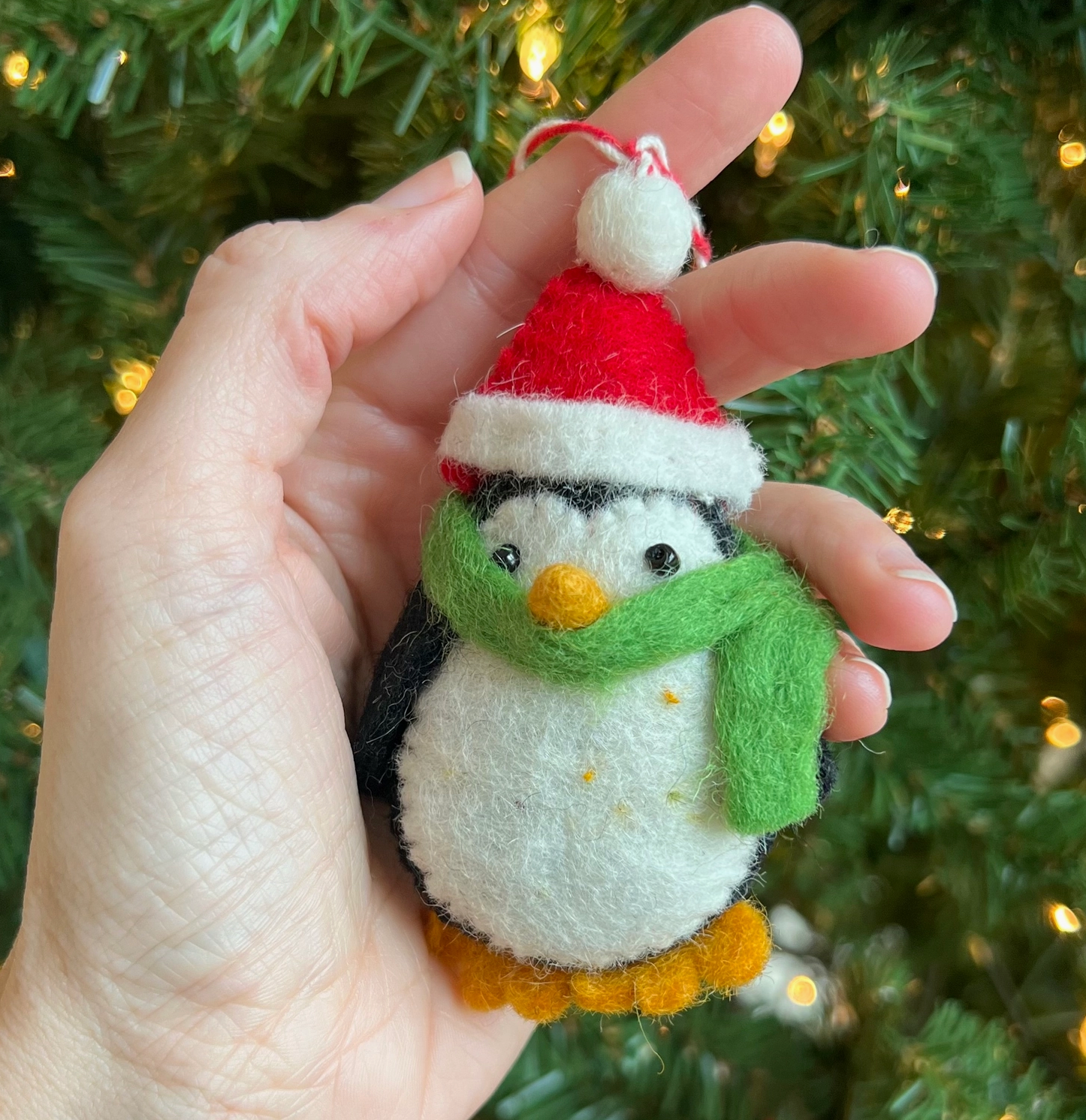 Handmade felt penguin Christmas ornament with red Santa hat, green scarf, and orange feet held in palm of hand