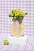 Yellow and pink checkered paper vase with yellow flowers on white pedestal, green apple beside it, purple gingham background