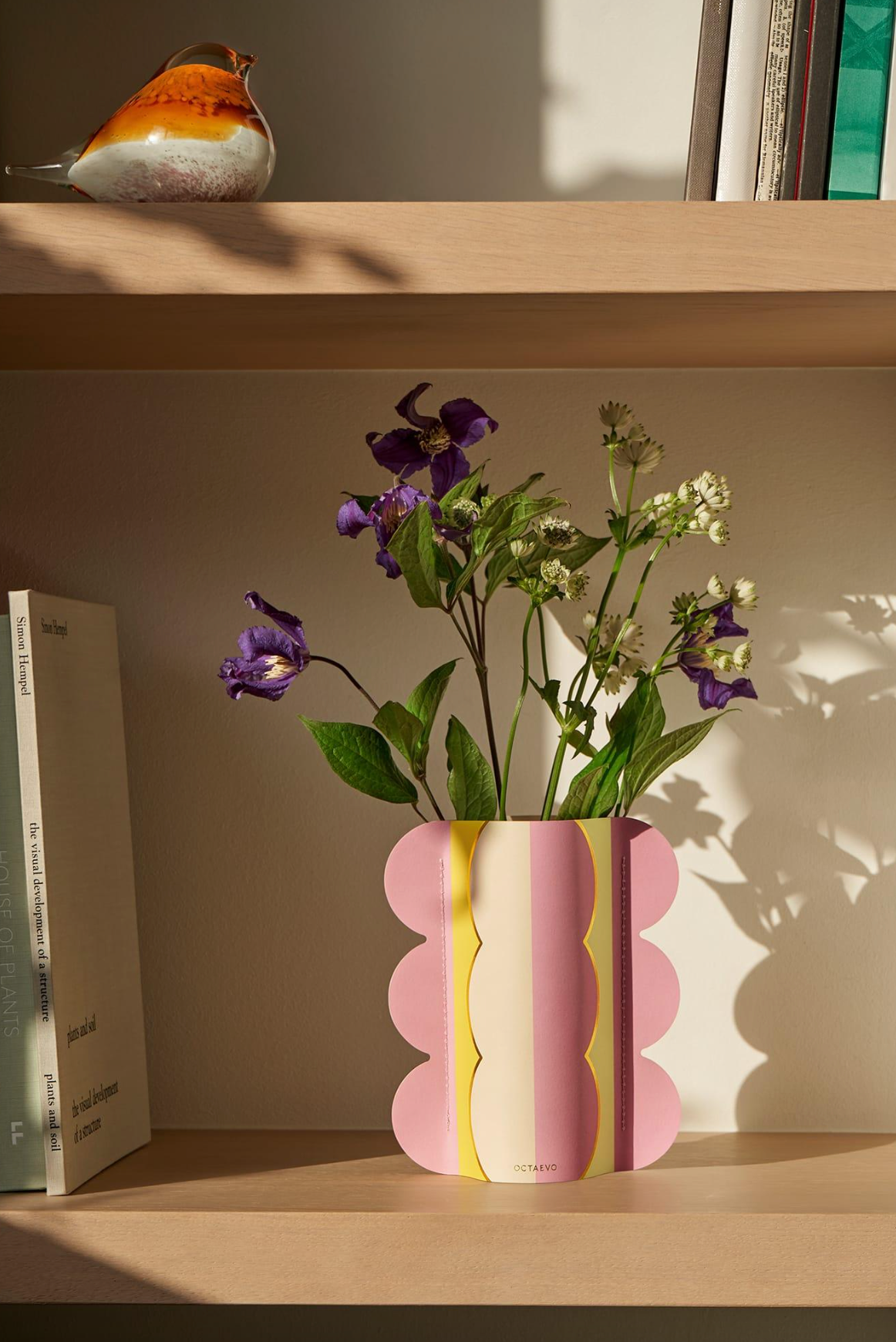 Pink ceramic vase with scalloped edges, yellow and cream vertical stripes, holding purple and white flowers on wooden shelf