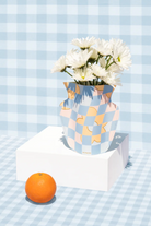 White daisies in a checkered paper vase with blue, peach, and yellow pattern, displayed on white platform beside an orange