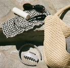Black and white gingham bikini with ruched detail displayed on sand with straw hat, sunglasses, and sunscreen bottle