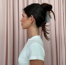Gray comb hair clip securing a high ponytail in dark brunette hair, worn by woman in white shirt against pink curtains