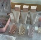 Three beige EMI JAY hair care tubes with matching boxes and claw clips displayed on marble surface with pink cosmetic bags