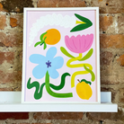Framed art print with colorful fruit and floral motifs including lemons, pink tulip, blue flower on pale pink background