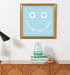 Framed blue wall art with white flower eyes and text "BLOOM WHERE YOU'RE PLANTED" forming a smile in a gold frame
