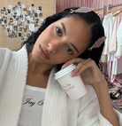 Woman wearing white fuzzy robe with black script text, holding white branded cup, pink headband in hair