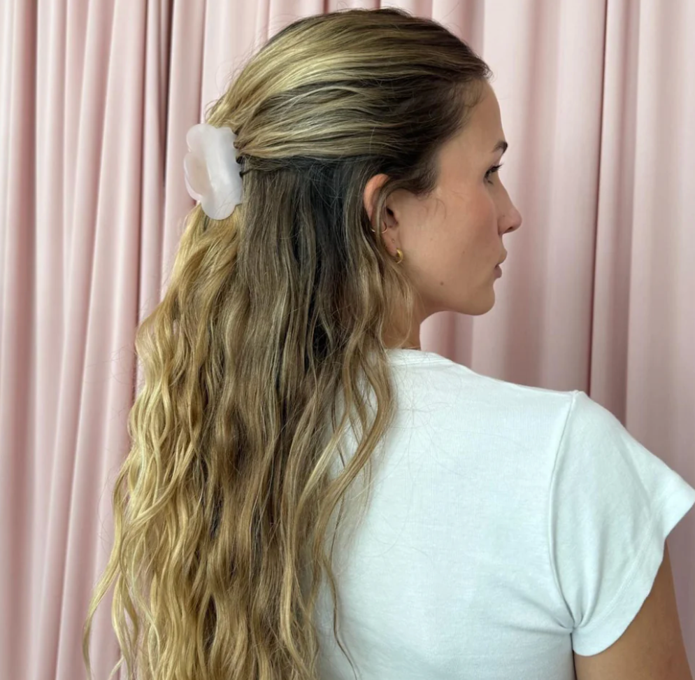White organza flower hair clip worn in wavy blonde hair in half-up hairstyle against pink curtain background