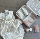 Pink satin cosmetic travel case with white products, gua sha tools, and comb displayed alongside white lace lingerie