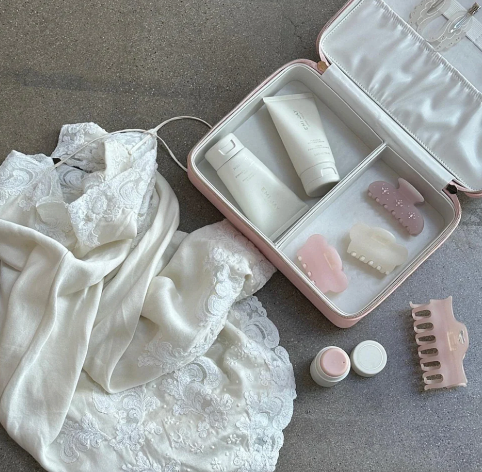 Pink satin cosmetic travel case with white products, gua sha tools, and comb displayed alongside white lace lingerie