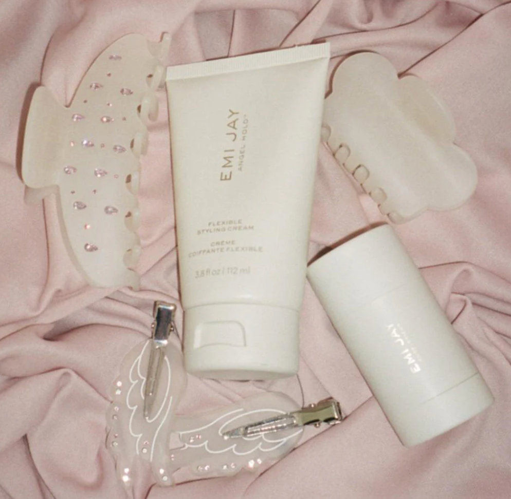 Beauty and skincare products including EMI JAY styling cream, gua sha tools, and facial roller on pink fabric background