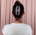 Large translucent gray resin hair claw clip holding dark hair in an updo bun against pink curtain backdrop