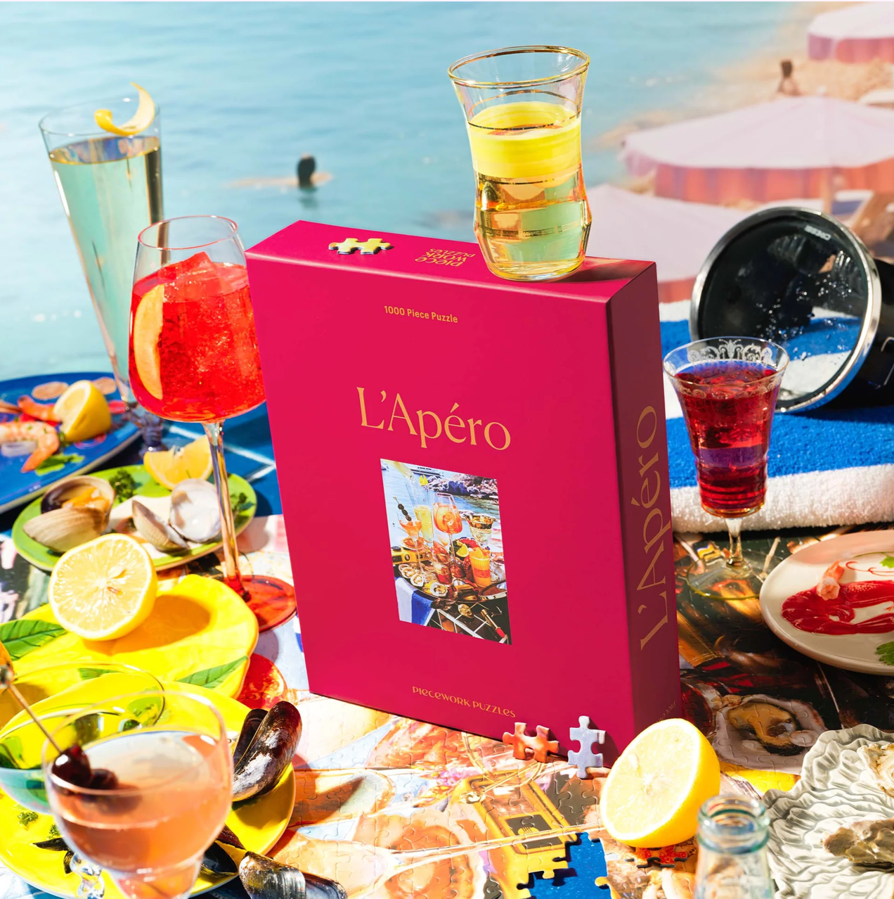 Bright pink puzzle box titled L'Apéro surrounded by colorful cocktails, citrus slices, and appetizers by a pool