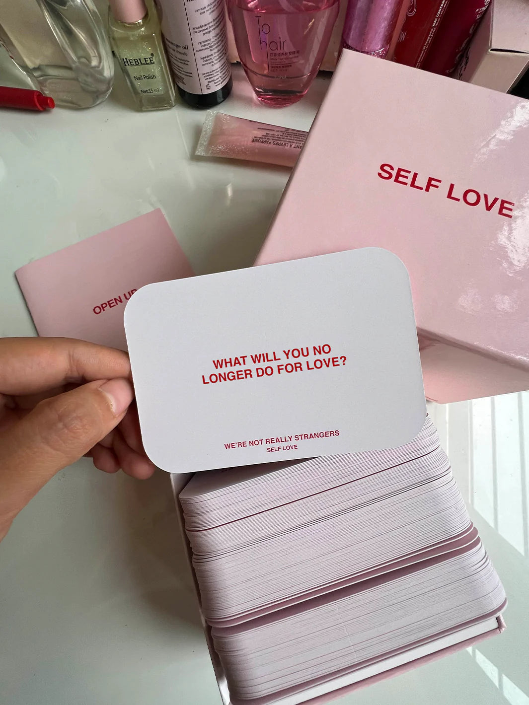 Hand holding white card with red text reading "What will you no longer do for love?" from self-love card deck on pink surface