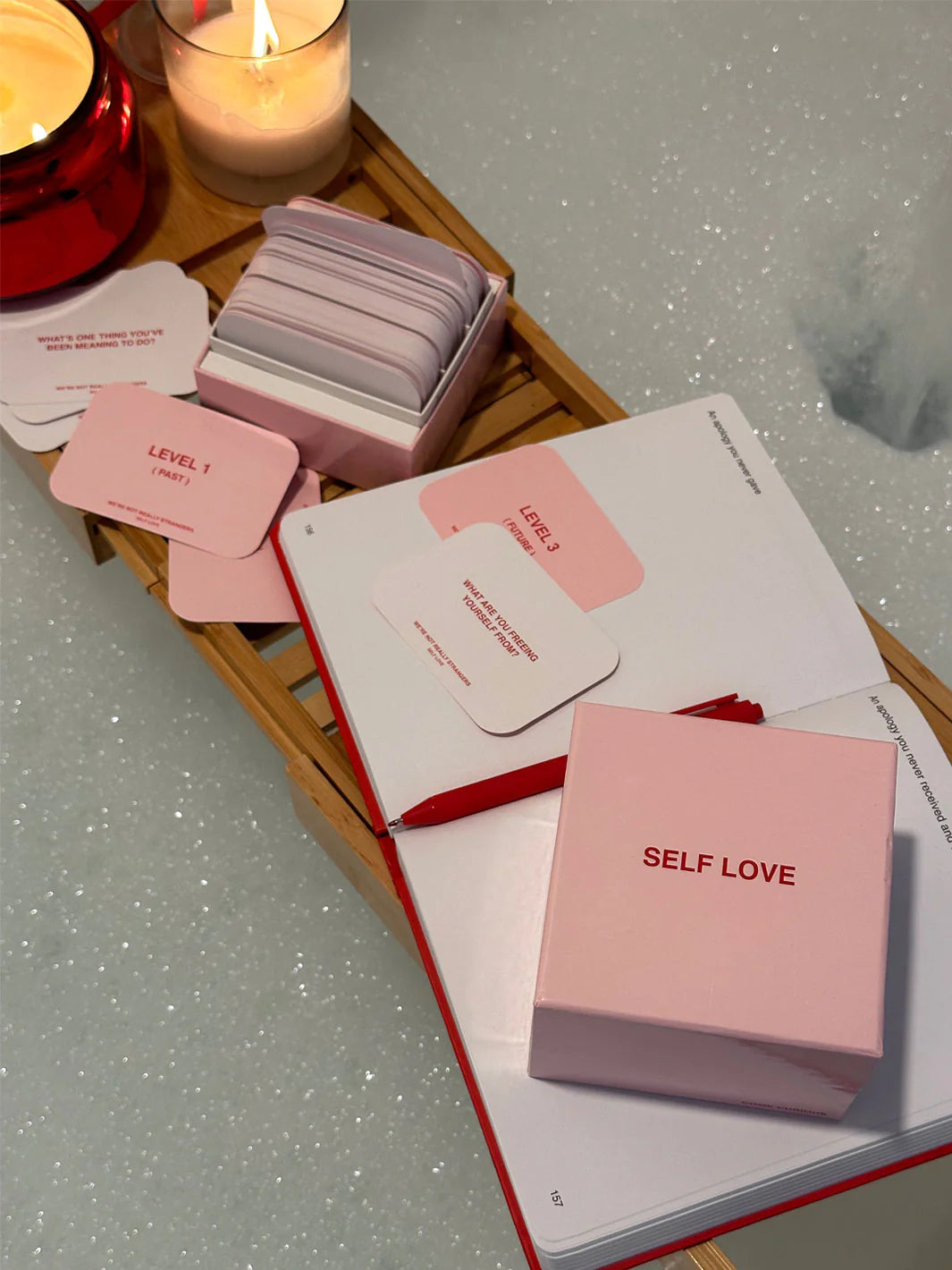 Pink self-love card deck with rounded corner cards in wooden tray beside candle and open journal on sparkly surface
