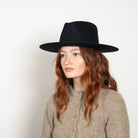 Black wide-brim fedora hat worn by woman with long wavy auburn hair and beige knit sweater against white background
