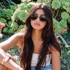 Woman wearing sunglasses sitting outdoors with greenery in the background