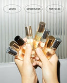 Hands holding multiple gender-neutral perfume bottles with gold and black caps against white background
