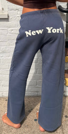 Navy sweatpants with 'New York' text worn by a person, against a stone wall background.