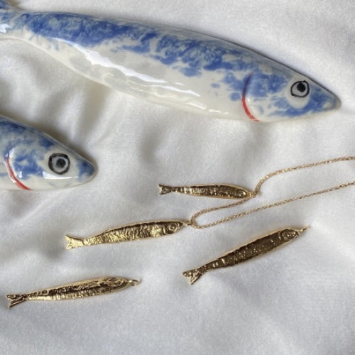 Gold fish pendant necklaces displayed on white fabric with blue and white ceramic fish decorations