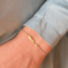 Delicate gold chain bracelet with rectangular bar pendant worn on wrist against light blue fabric sleeve
