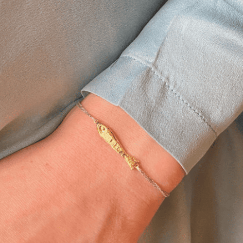 Delicate gold chain bracelet with rectangular bar pendant worn on wrist against light blue fabric sleeve