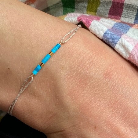 Delicate silver chain bracelet with turquoise blue beaded bar accent on wrist against colorful plaid fabric background