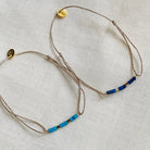 Two delicate adjustable bracelets with beige cord and blue beaded accents, one in light blue and one in navy blue