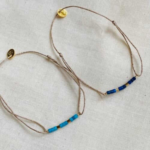 Two delicate adjustable bracelets with beige cord and blue beaded accents, one in light blue and one in navy blue