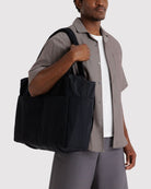 Black canvas tote bag with front pocket carried over shoulder by person wearing gray shirt and pants