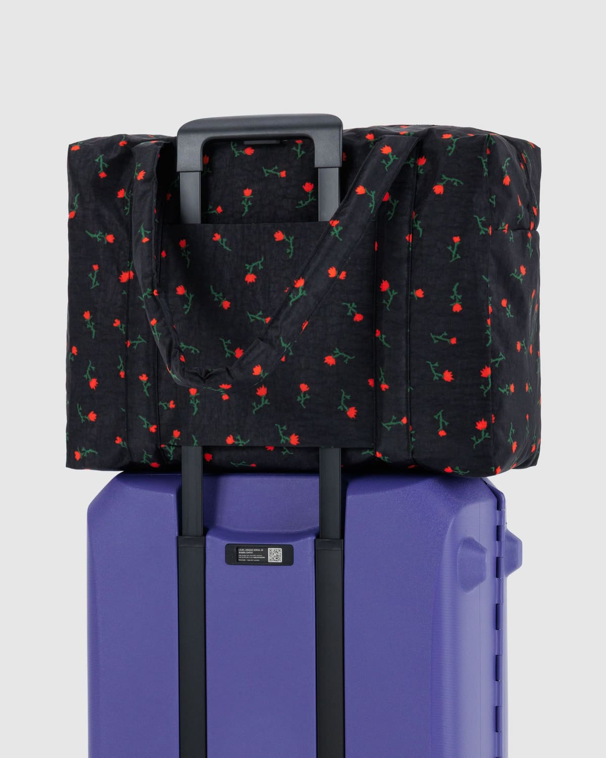 Black duffel bag with red rose print pattern attached to purple rolling suitcase via luggage sleeve