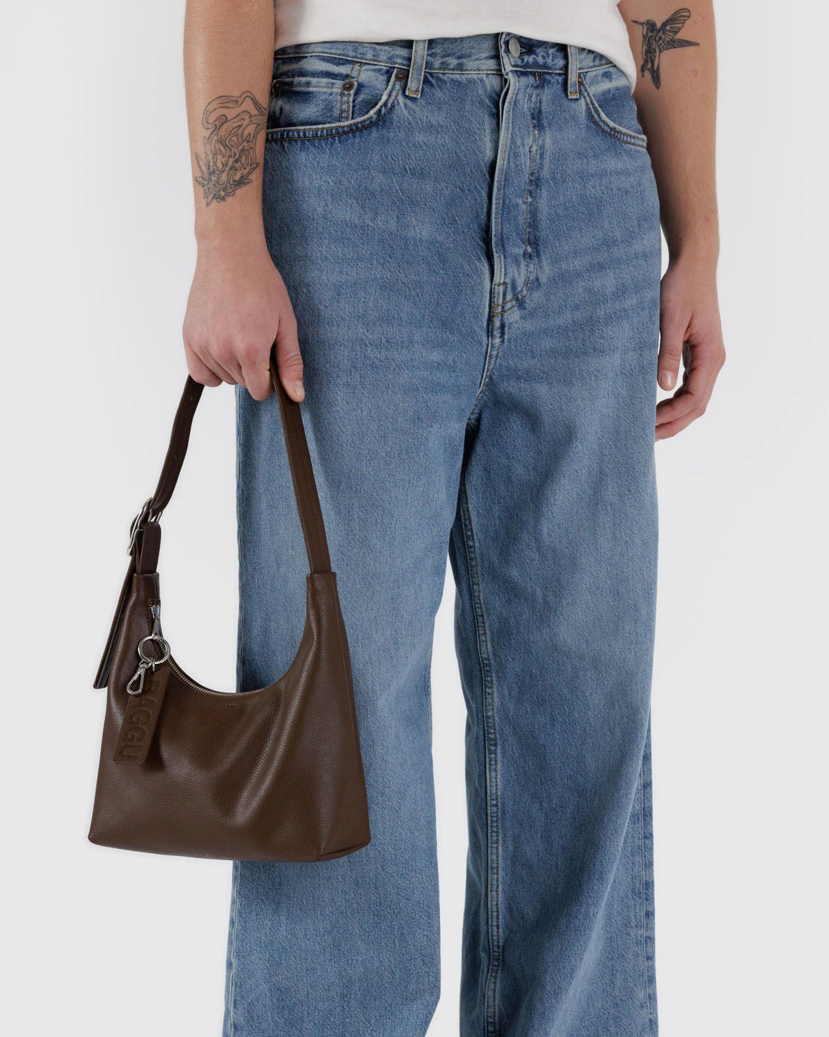 Brown leather hobo shoulder bag with dual straps and keychain charm held against light-wash denim jeans