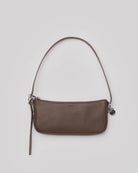Brown leather shoulder bag with slim strap, zip closure, and silver hardware charm on neutral background