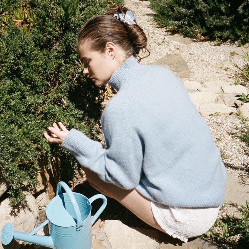 Woman wearing oversized light blue knit sweater with ribbed collar, crouched outdoors near blue watering can