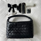 Black woven leather shoulder bag displayed with makeup accessories including brushes, bow clip, eyelash curler and nail clippers on white fabric