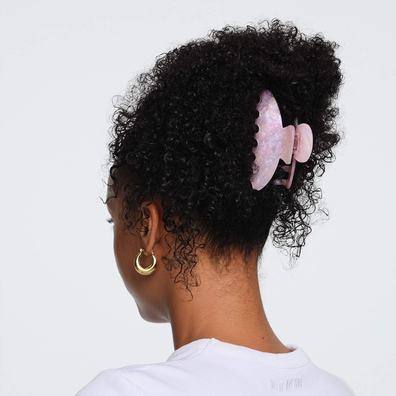 Rose quartz gua sha tool clipped in curly black hair, shown from behind on model wearing gold hoop earring
