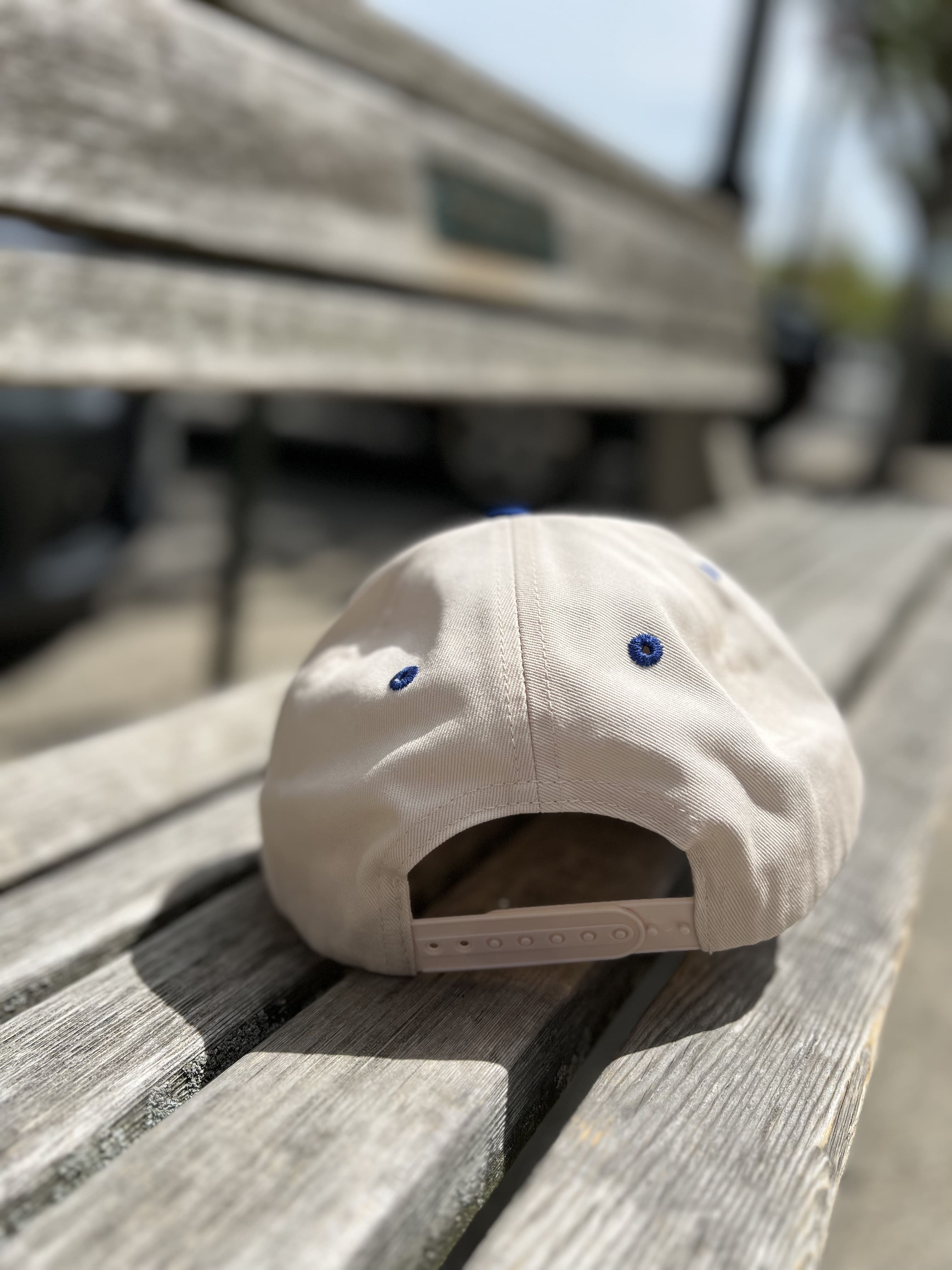White snapback hat with blue eyelets resting on wooden dock surface with boat in background