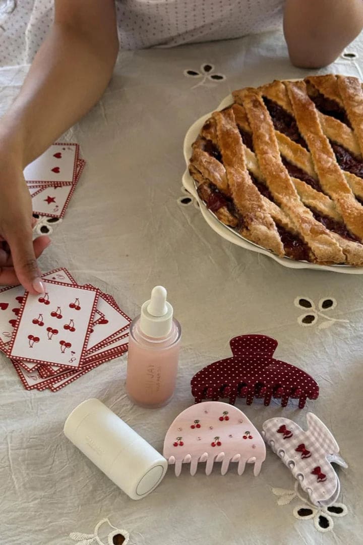 Cherry-patterned hair accessories set including pink claw clips, hair ties, and styling tools arranged on white fabric with pie