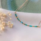 Delicate gold chain necklace with turquoise blue beads in curved design, displayed on white fabric with dried flowers
