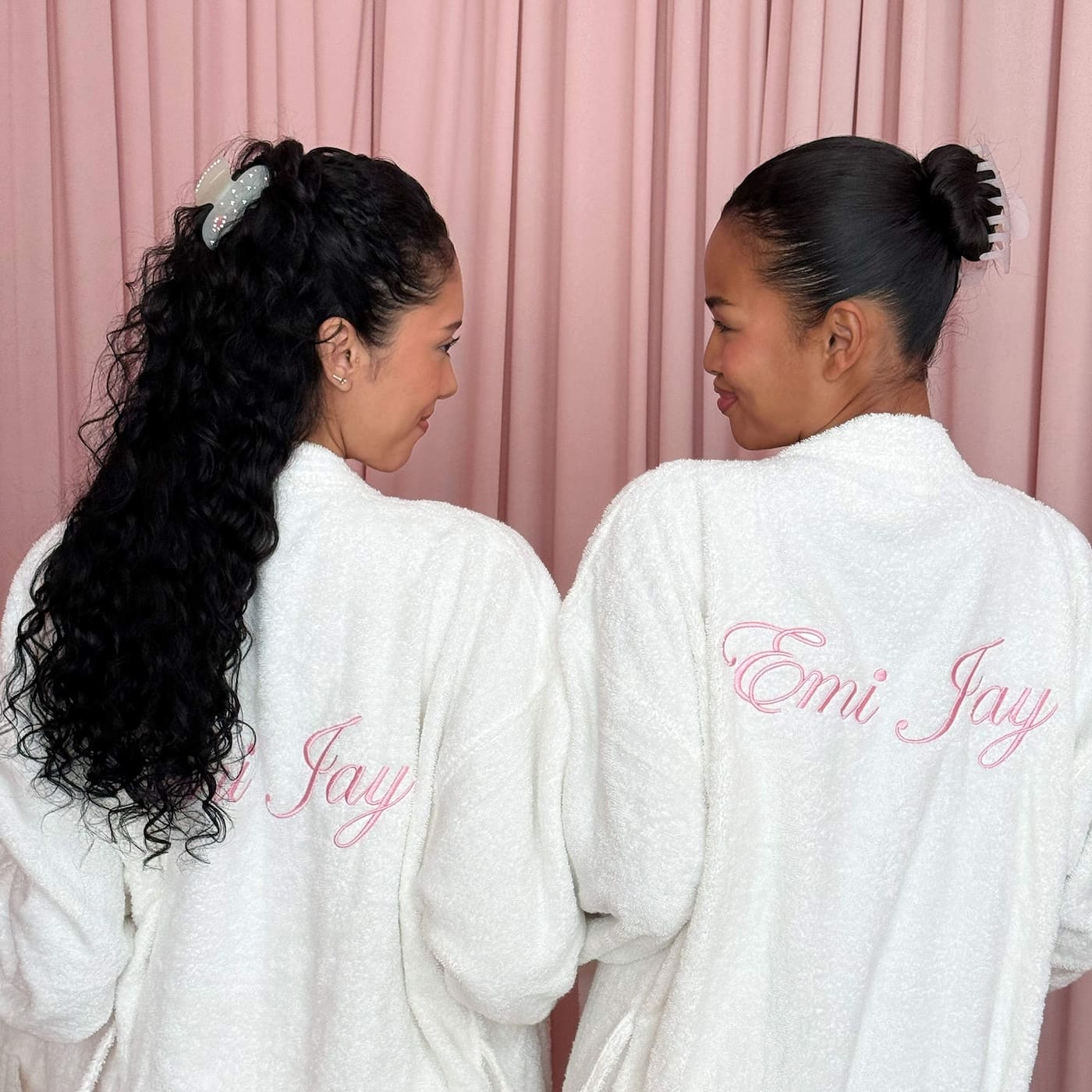 Two women wearing white plush robes with pink embroidered "Emi Jay" text on back, showing different hairstyles with matching scrunchies