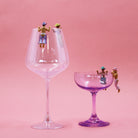Two glasses, one large and one small, with miniature figures on a pink background