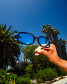Black sunglasses with blue reflective lenses held up against palm trees and clear blue sky