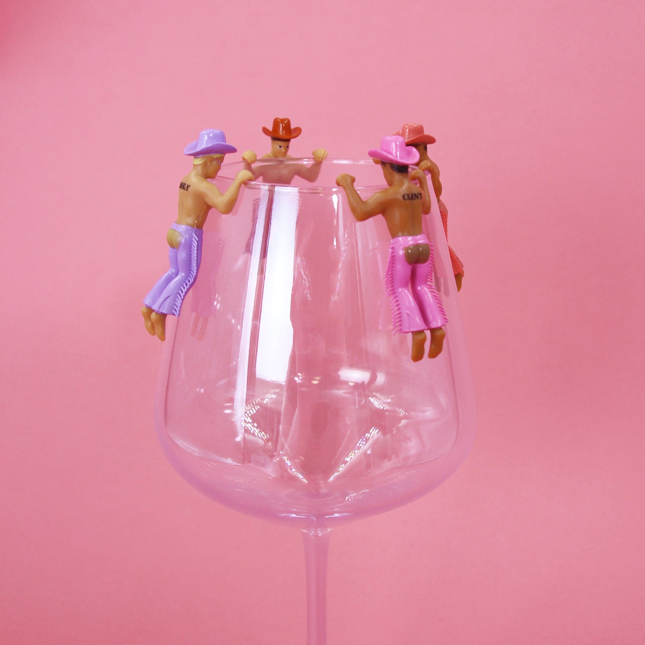 Three miniature cowboy figurines in purple and pink outfits positioned around the rim of a clear wine glass on pink background