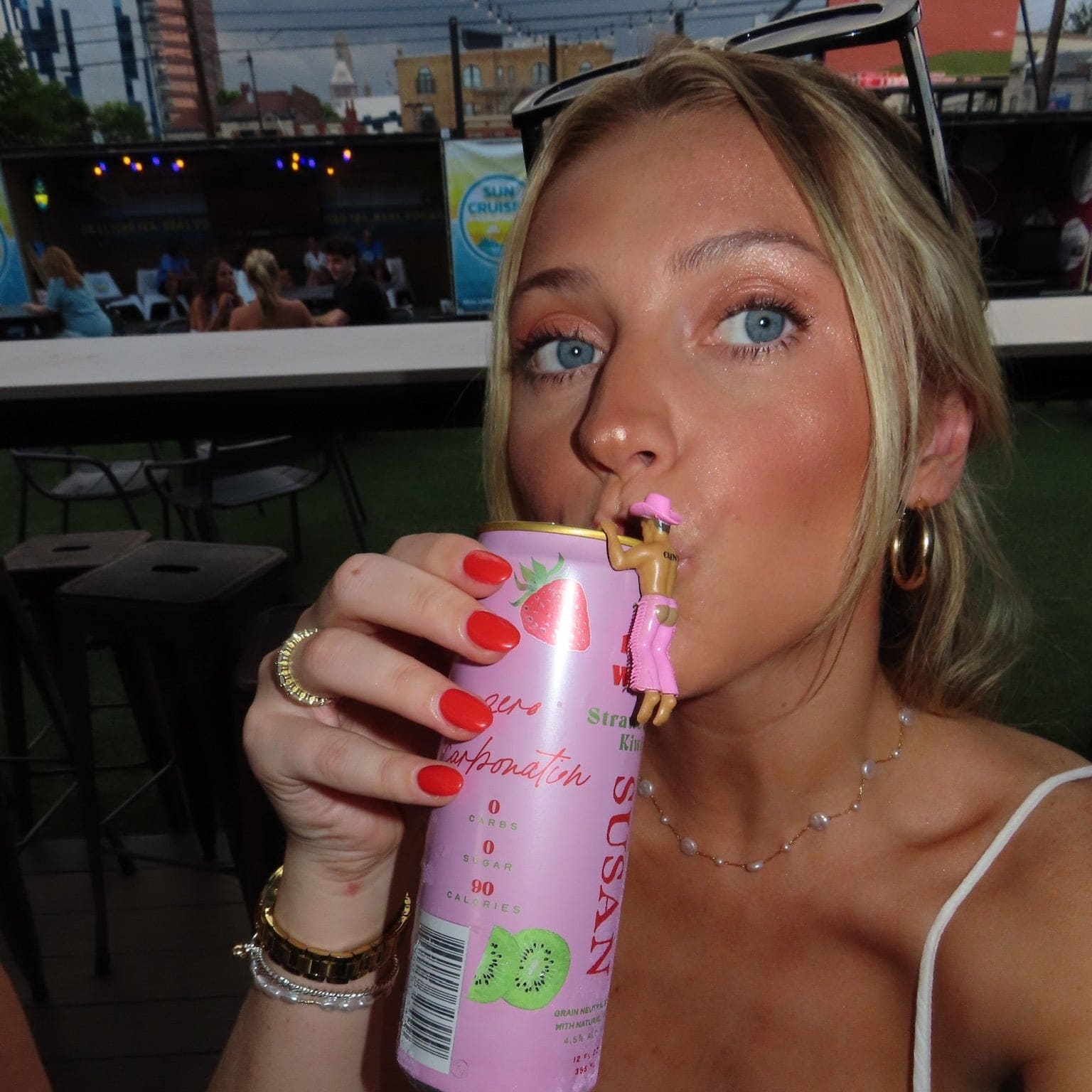 Woman drinking from pink can with strawberry and kiwi fruit graphics, wearing white tank top and jewelry at outdoor venue